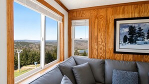 A room with a gray leather sofa, large windows with wooden frames, and a framed picture of a snowy landscape. - ML 342: Breathtaking Views & Prime Location for the Perfect Summer Stay (Snowshoe)