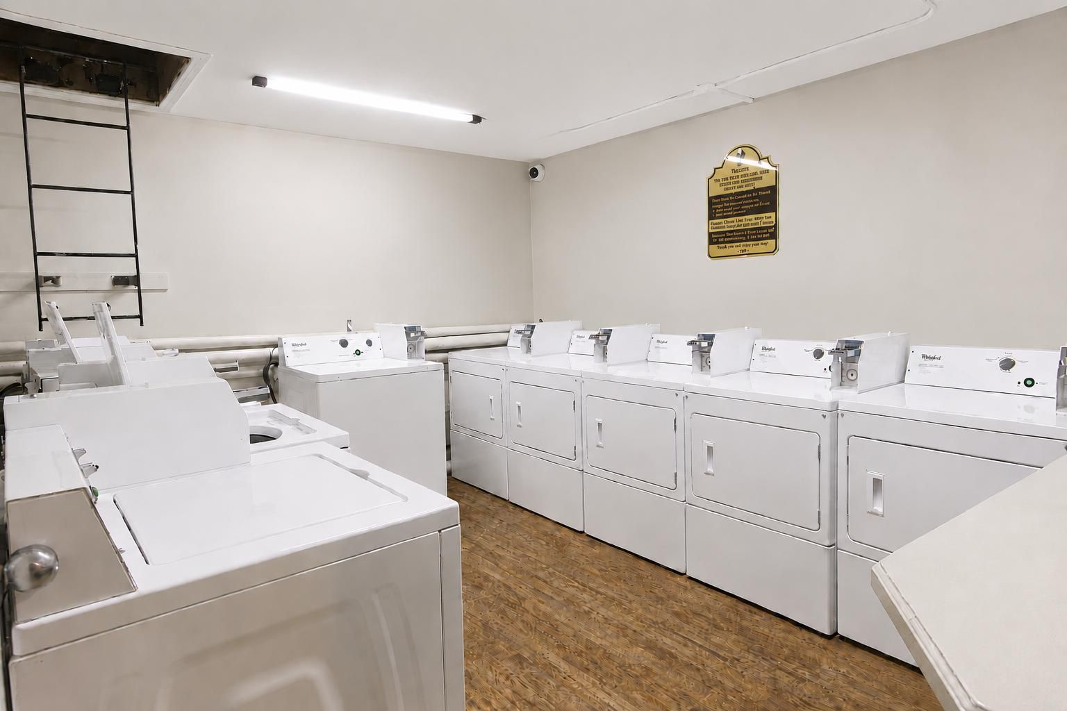 A laundry facility with multiple washing machines and dryers, a wooden floor, and a wall-mounted plaque.