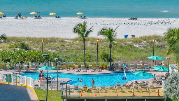 A pool area with lounge chairs and umbrellas, a beach with people and birds, and a clear view of the ocean. - AMAZING BEACH VIEW/ FULLY REMODELED CONDO/ALL NEW FURNITURE/ HEATED POOL (Treasure Island)