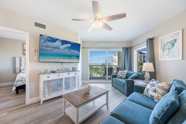 A living room with a ceiling fan, a large TV, a sofa, a coffee table, and a view of the outside.