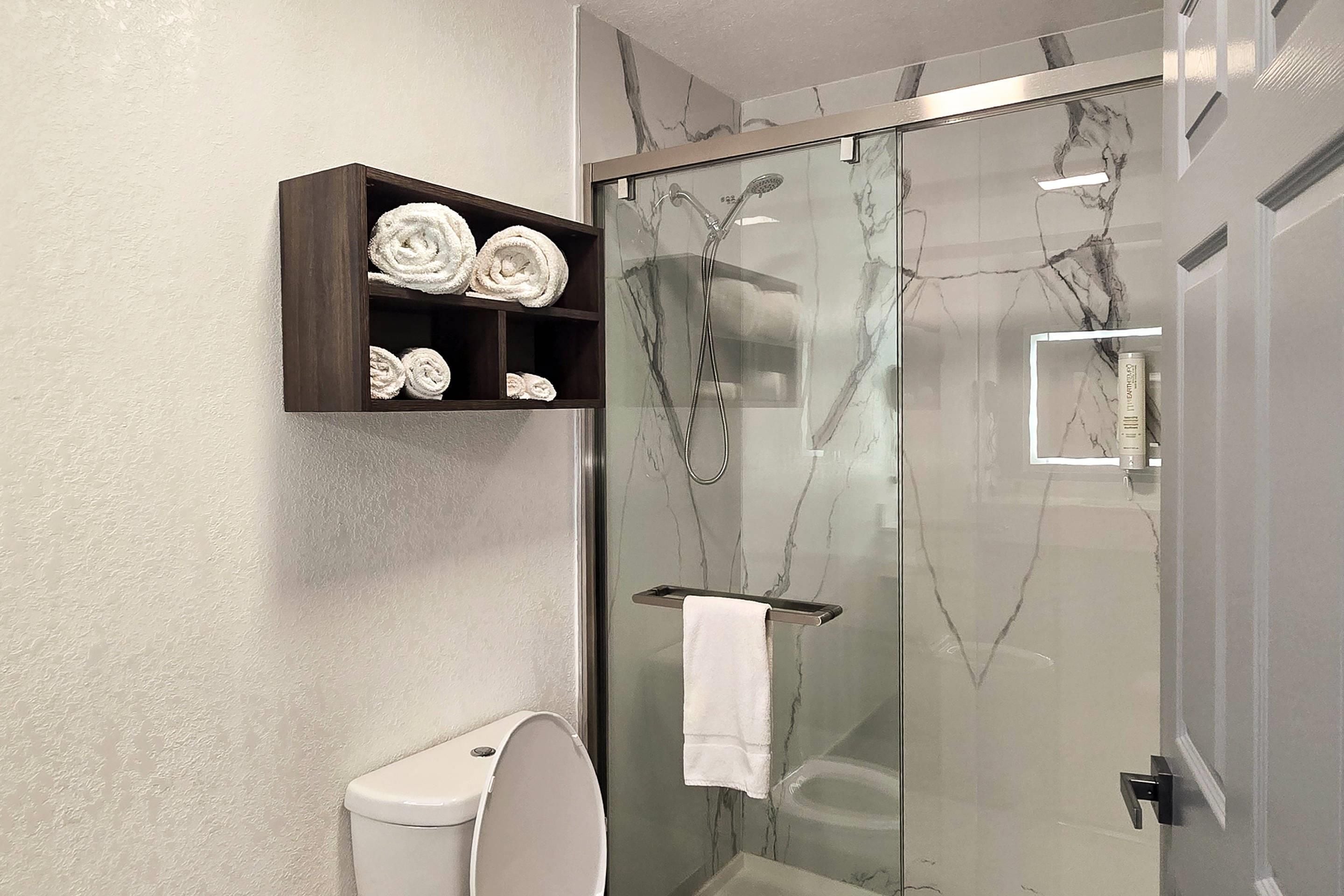 A bathroom with a glass-enclosed shower, a toilet, and a shelf with rolled towels.