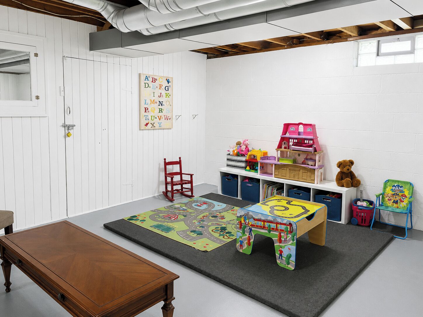 A children's playroom with a wooden table, play mats, a bookshelf, and toys.