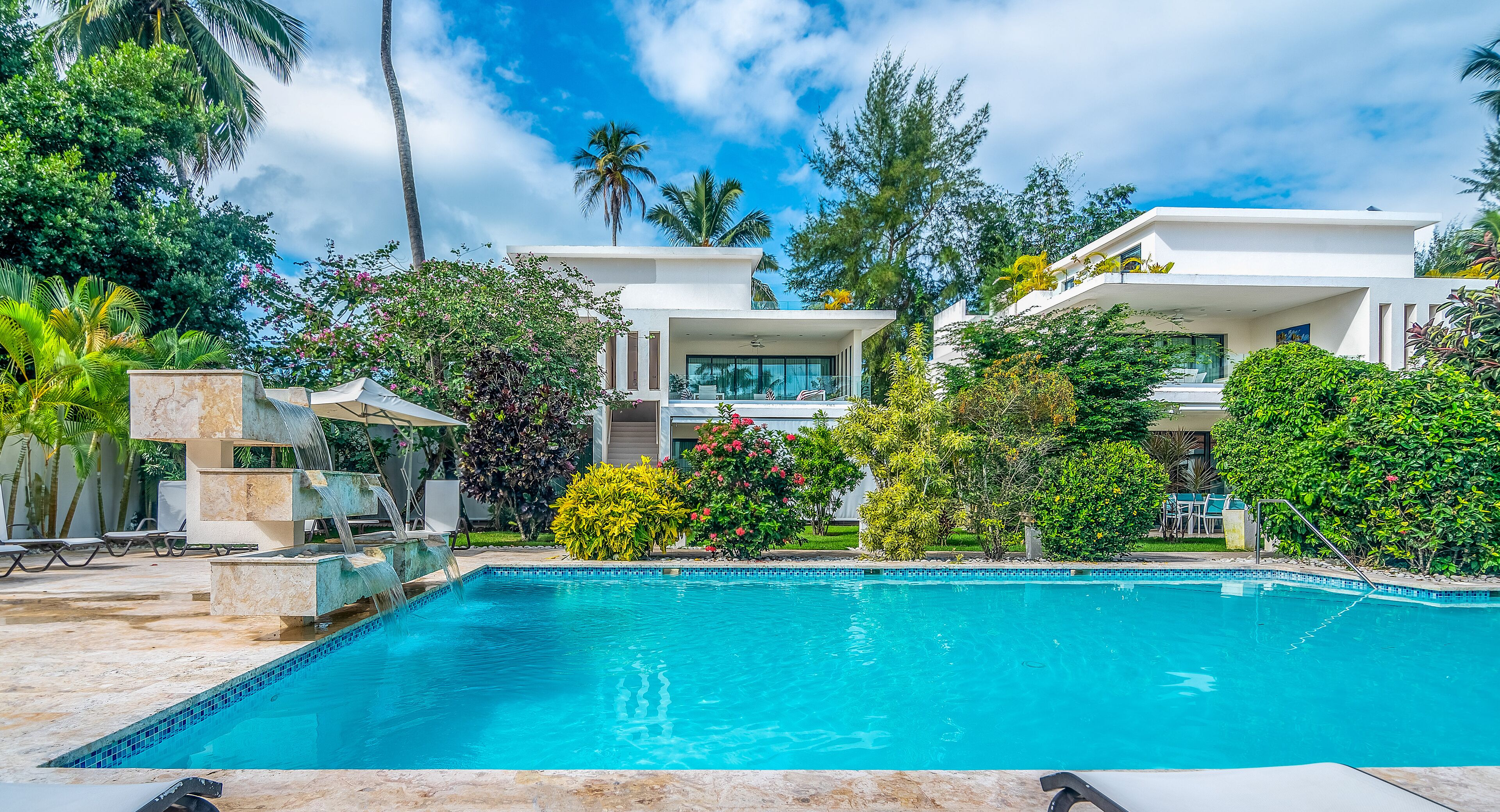 A modern pool area with a clear blue pool, surrounded by lush greenery and white buildings.