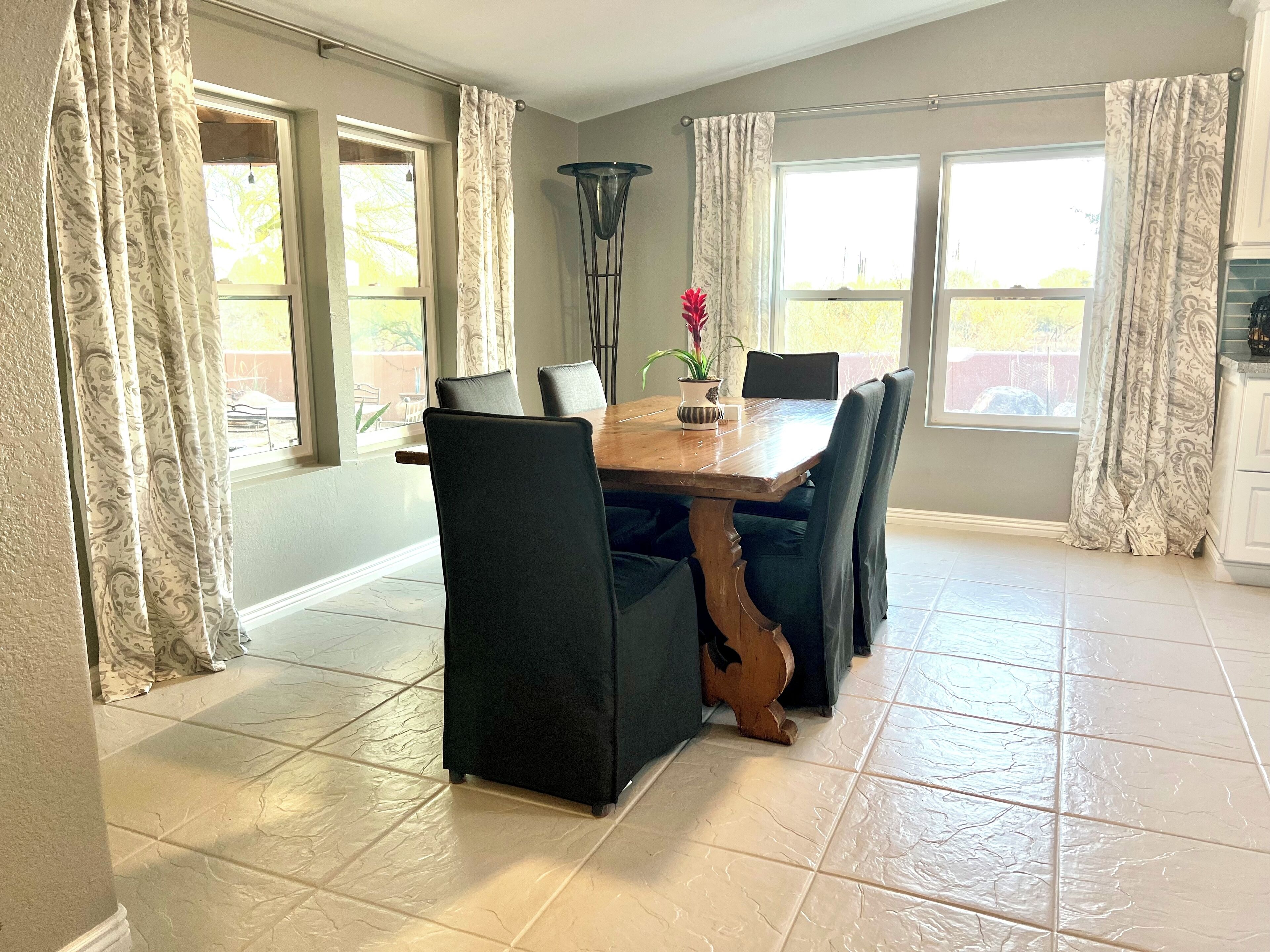 Un espace repas avec une table en bois et des chaises noires, de grandes fenêtres avec des rideaux et un sol carrelé.