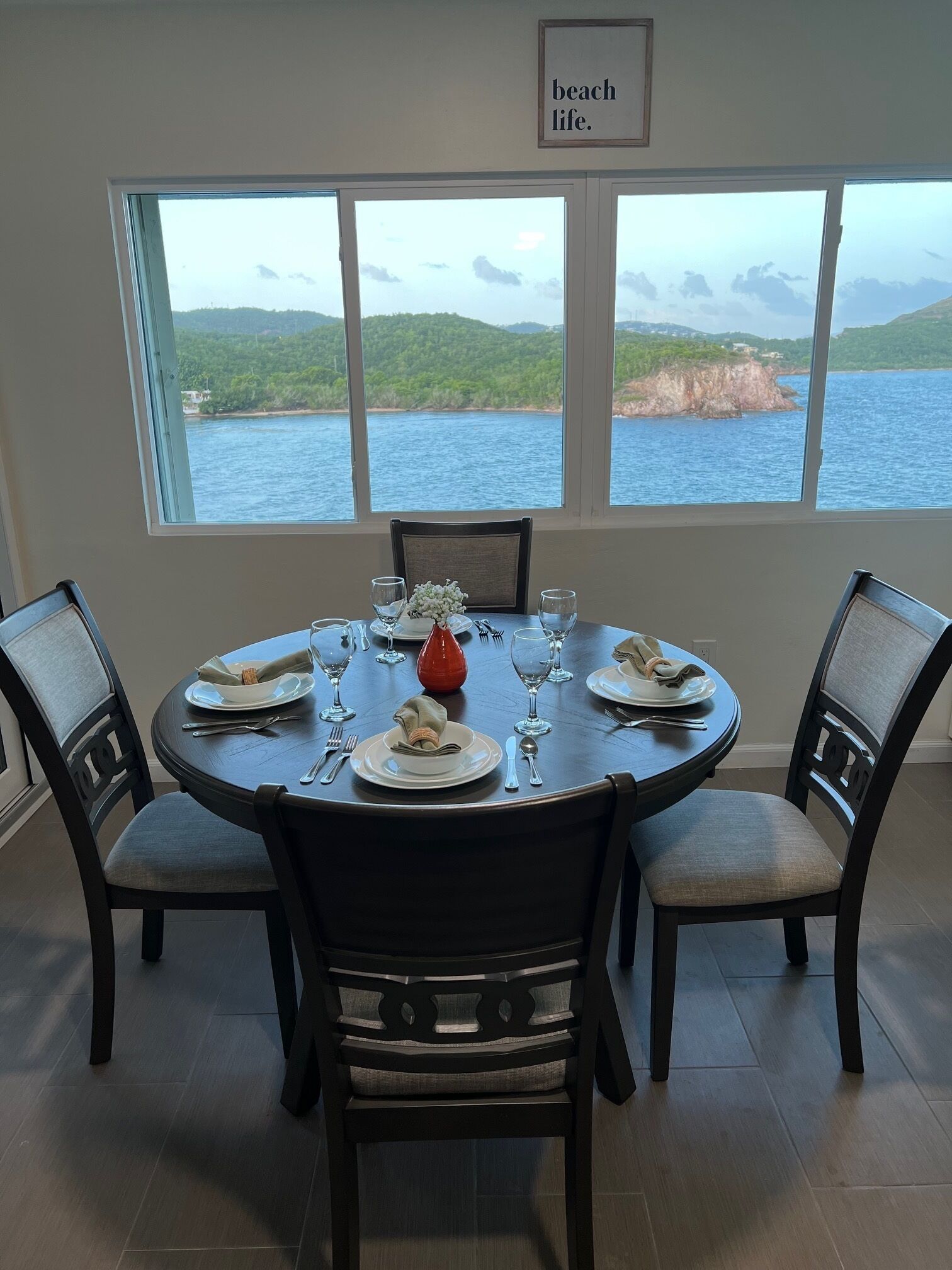 A dining area with a round table set for four, large windows offering a view of a body of water and hills, and a framed sign above the windows reading "beach life."