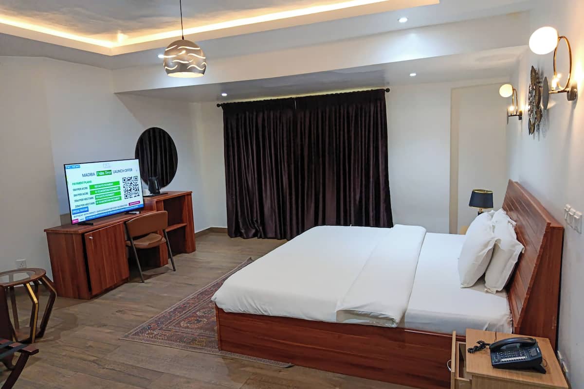 Une chambre d’hôtel avec un lit en bois, un bureau avec un ordinateur, une chaise, un téléphone et un téléviseur diffusant une publicité.