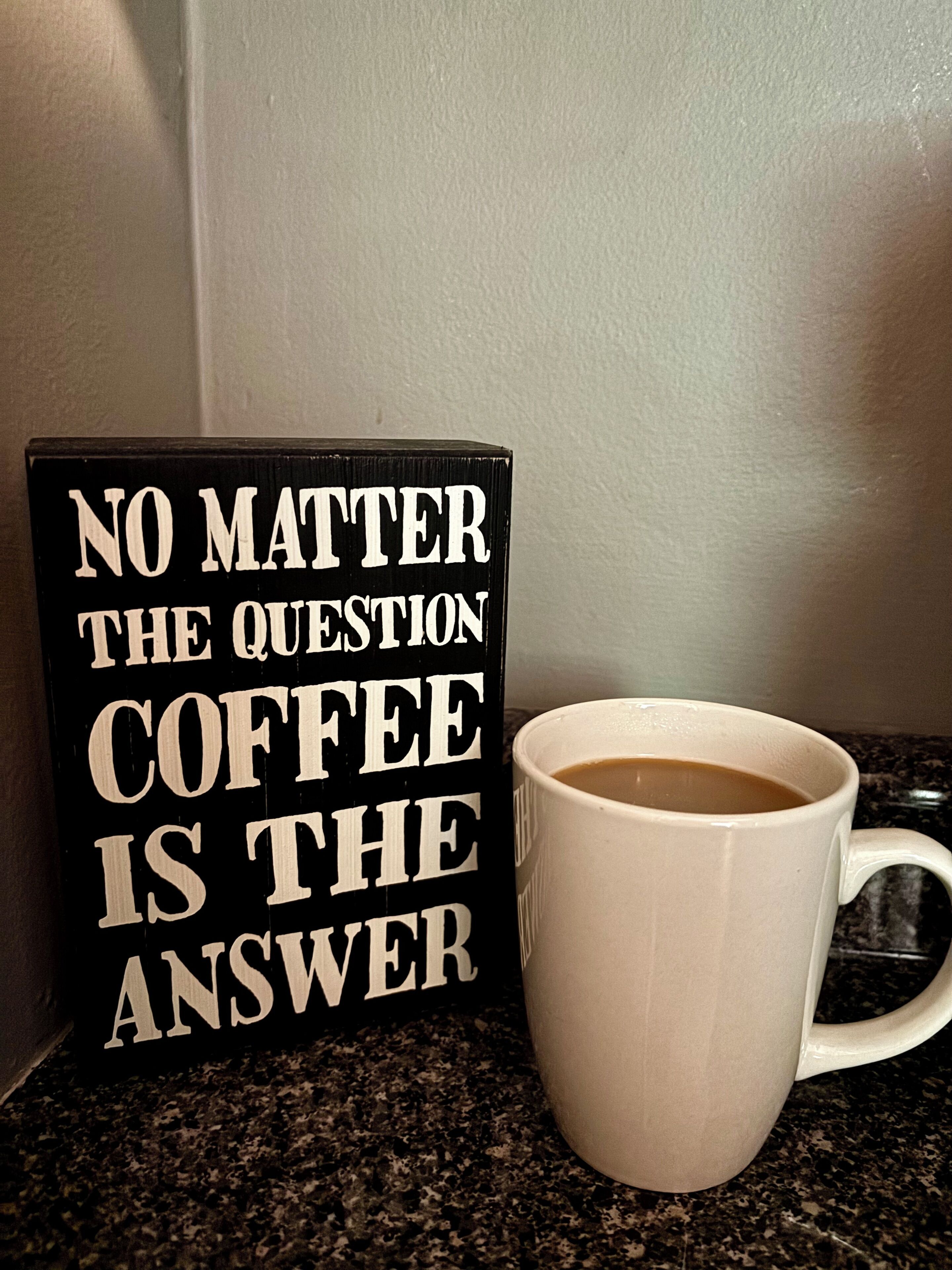 A black sign with white text and a white mug with coffee on a granite countertop.