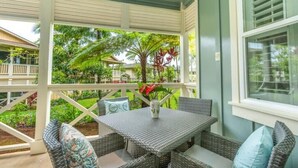 A covered outdoor seating area with wicker furniture and a table, surrounded by lush greenery and a white railing. - Princeville Paradise - Luxury A/C (Princeville)