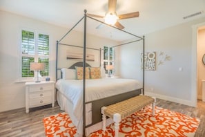 Un dormitorio con cama con dosel, ventilador de techo, mesita de noche con lámpara y ventana con persianas.