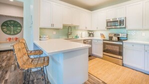 A kitchen with white cabinets, a microwave, an oven, and a sink. There is a clock on the wall and a woven chair. - Princeville Paradise - Luxury A/C (Princeville)