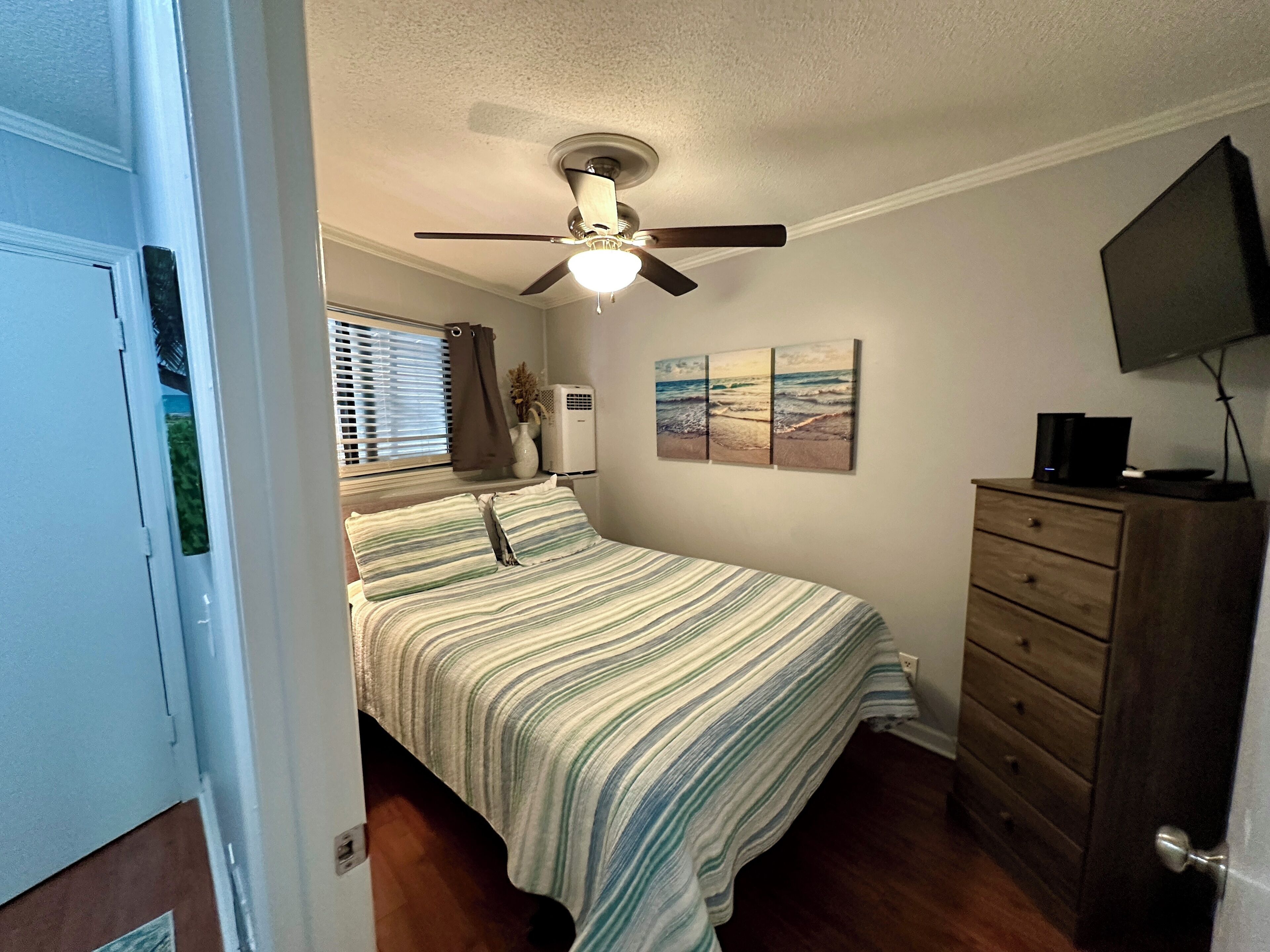A bedroom with a bed, a ceiling fan, a dresser, a TV, and artwork on the wall.