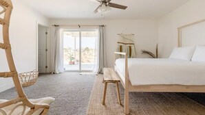 A bedroom with a bed, a wooden chair, a small table, a ceiling fan, and a view of the outdoors through a sliding door.