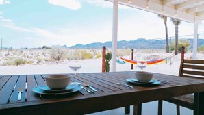 A wooden table set for two with bowls and a wine glass, overlooking a desert landscape with mountains in the distance.