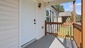 A covered porch with a wooden railing, a white door, and a small shed in the background.