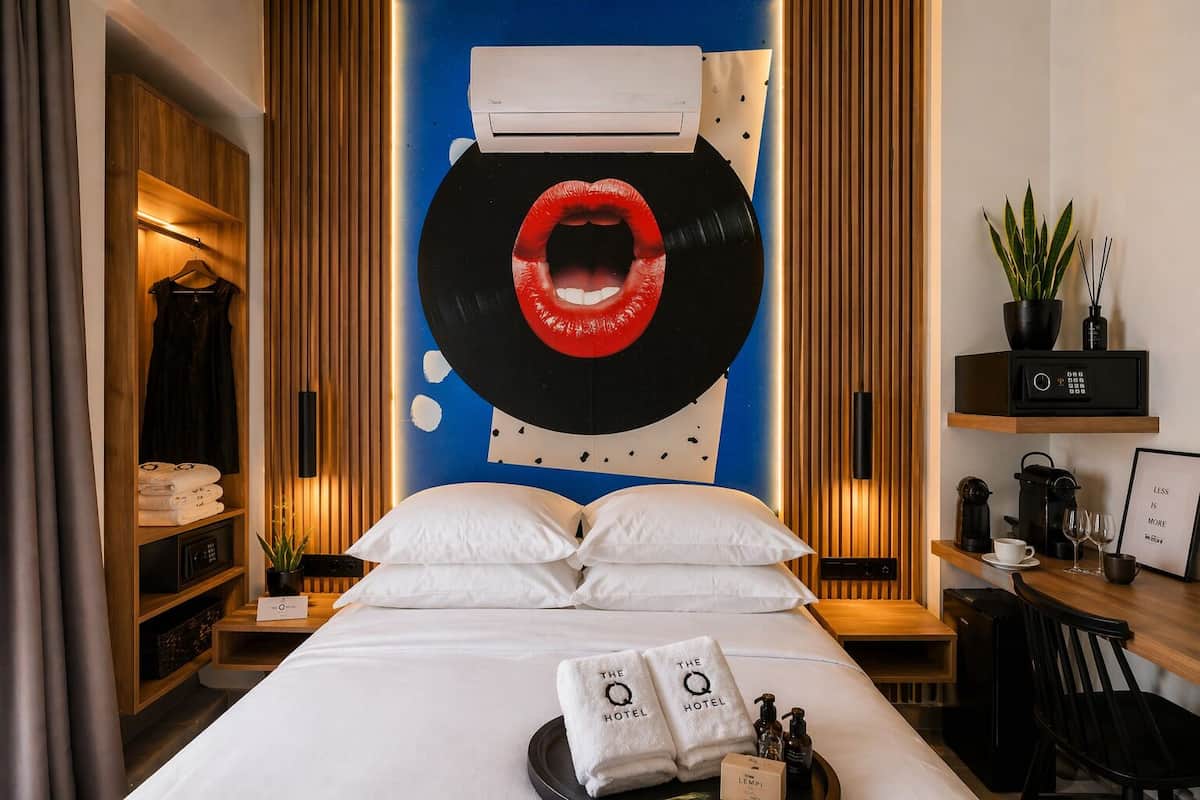 A hotel room with a bed, a wall-mounted air conditioner, a shelf with toiletries, and a decorative wall piece.