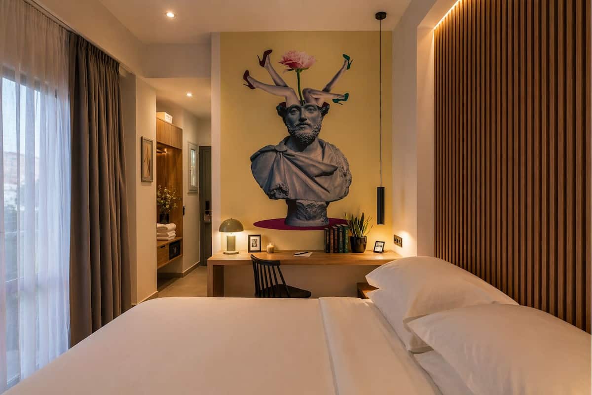 A hotel room with a large bed, a desk with a lamp, a bust sculpture on the wall, and a view of the city through the window.