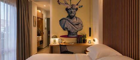 A hotel room with a large bed, a desk with a lamp, a bust sculpture on the wall, and a view of the city through the window.