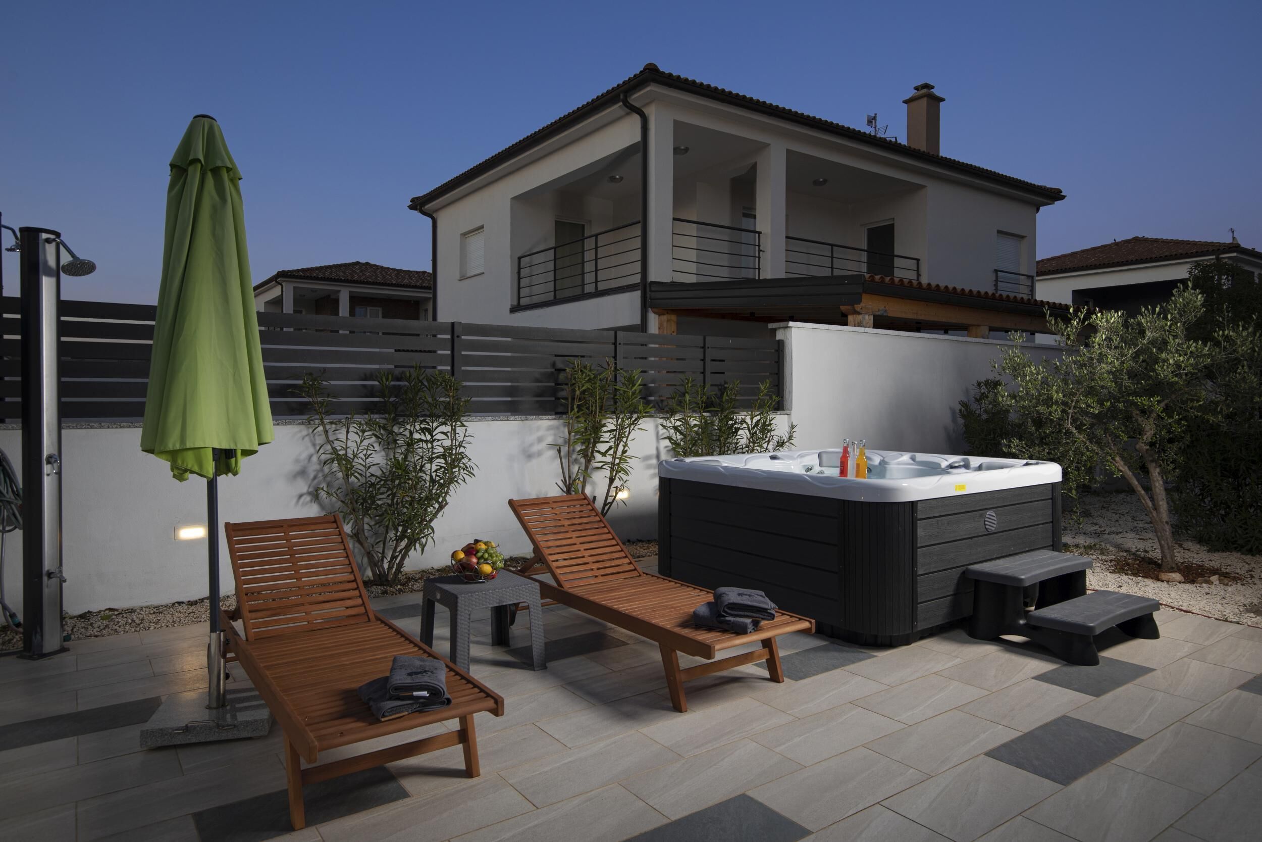 An outdoor patio with two wooden lounge chairs, a green umbrella, a hot tub, and a small table with fruit.