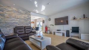 A modern living room with a stone wall, leather sofas, a flat-screen TV, and a dining area in the background.
