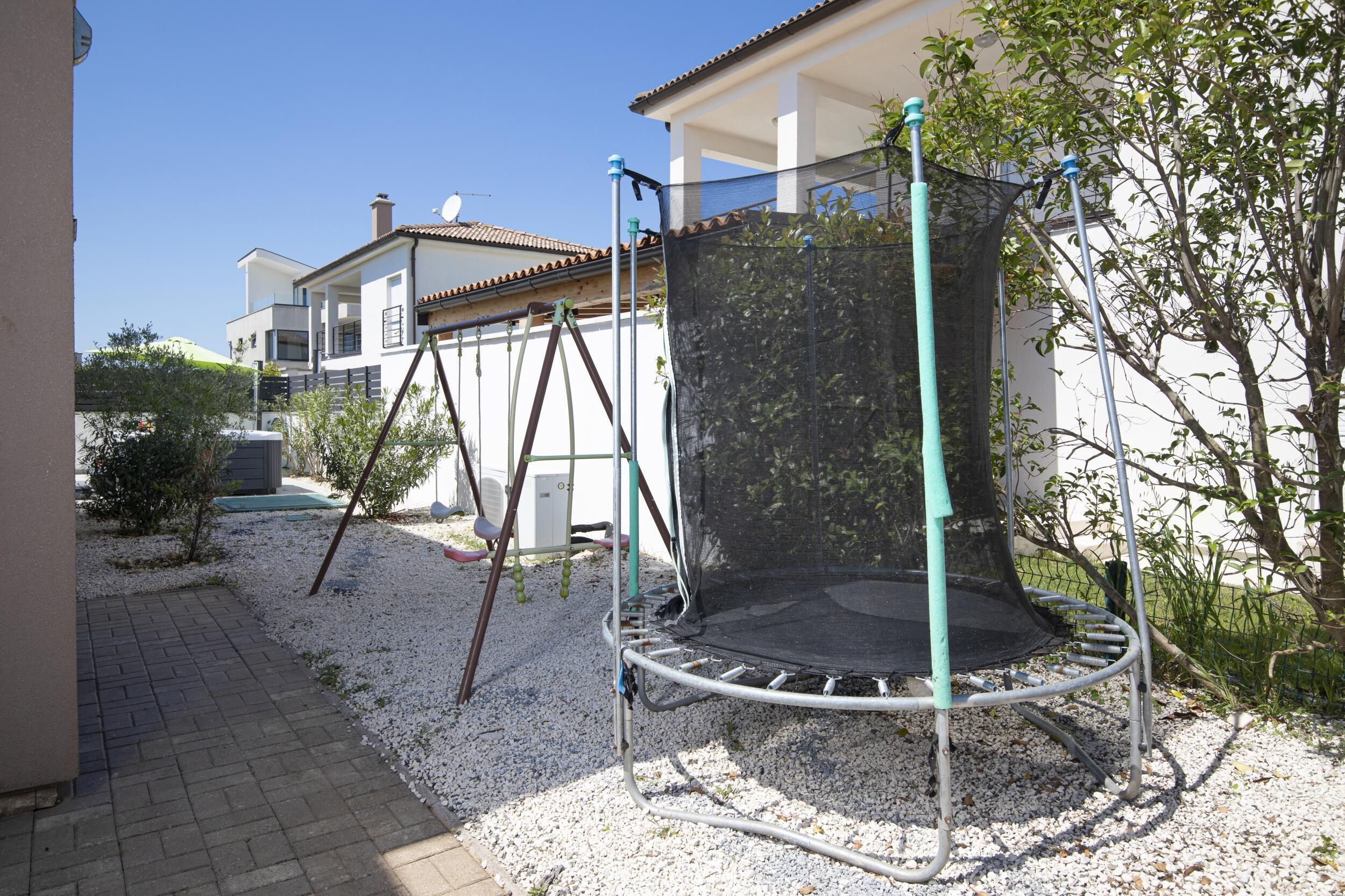 A trampoline in a backyard with a white fence, a brick pathway, and a house in the background.