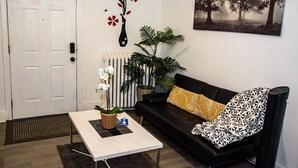 A living room with a white door, a black sofa with patterned cushions, a white coffee table, a potted plant, and a framed picture on the wall. - Modern Private Share Bedroom Bus stop to NYC (Englewood)