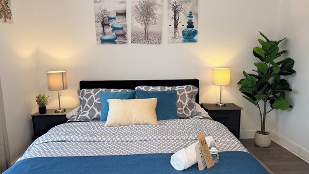 A neatly made bed with patterned bedding, two bedside tables with lamps, a large green plant, and three wall art pieces above the bed. - Modern Private Share Bedroom Bus stop to NYC (Englewood)