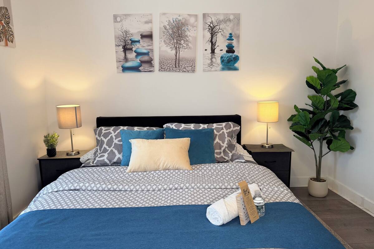 Cama bem arrumada com roupa de cama estampada, duas mesas de cabeceira com luminárias, uma planta verde grande e três quadros na parede acima da cama.
