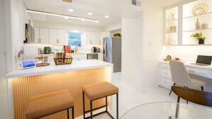Una cocina moderna con armarios blancos, una isla central con taburetes, un refrigerador y un comedor con mesa y sillas de vidrio.