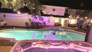 Una piscina con un jacuzzi al frente, rodeada de tumbonas e iluminada con luces colgantes.