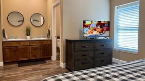 Une chambre à coucher comprenant un lit, une commode, un téléviseur et une fenêtre avec des stores.