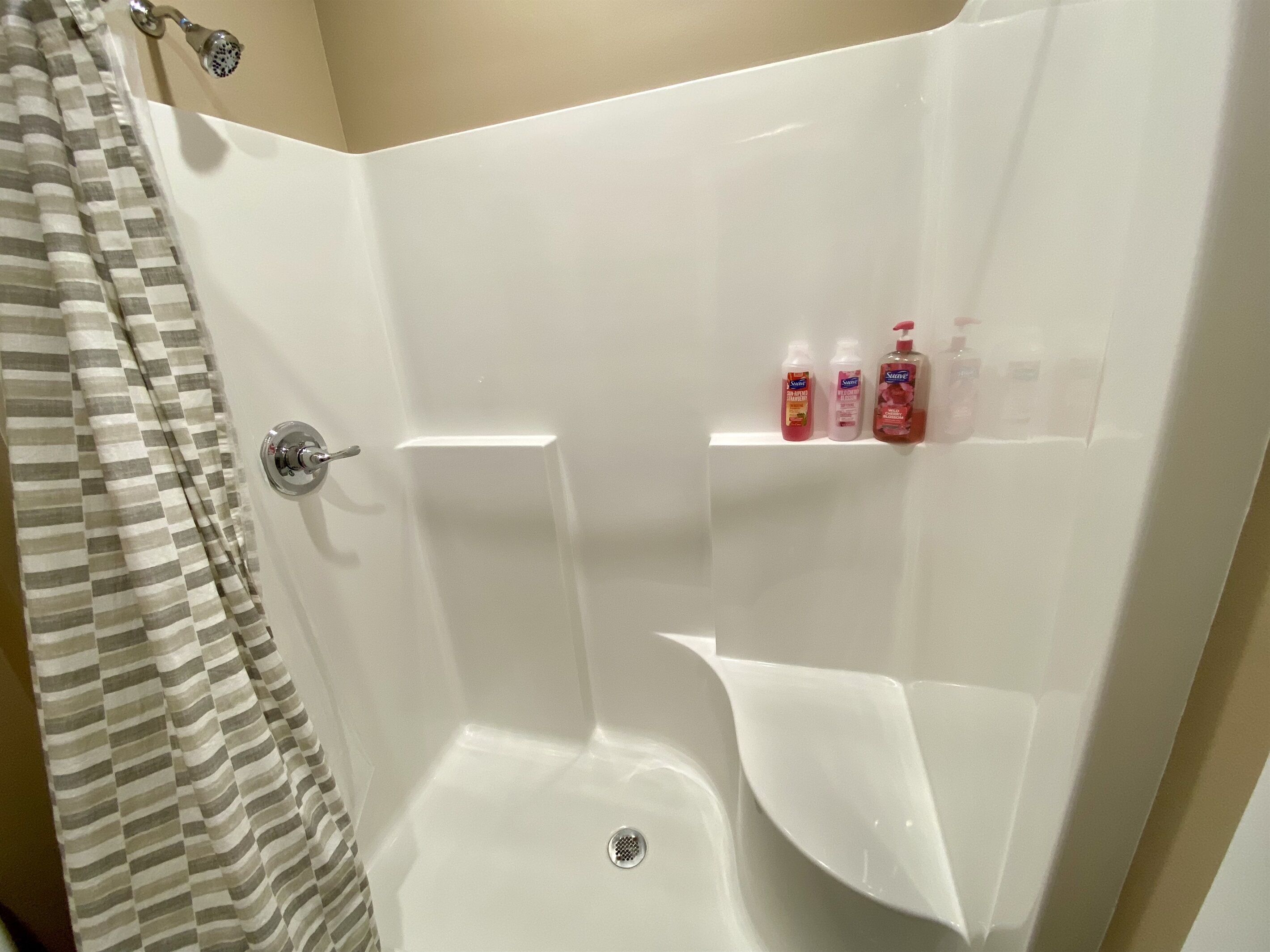 Ducha con pared de azulejos blancos, cabezal de ducha y estante con tres botellas de champú.