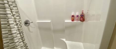 Ducha con pared de azulejos blancos, cabezal de ducha y estante con tres botellas de champú.