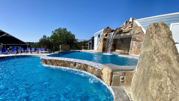 A swimming pool with a waterfall feature, surrounded by stone walls and blue chairs. - ELITE📍nearSIGHTnSOUND 🏊‍♀️POOL/WaterFall💦HOTTUB ⚡EV 🔥FIREPIT 🎱POOLTABLE 🎡 (Branson)