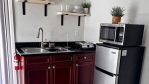 A modern kitchen with dark red cabinets, a stainless steel refrigerator, a microwave, and a gas stove. There are two shelves above the sink holding cups and a small plant.