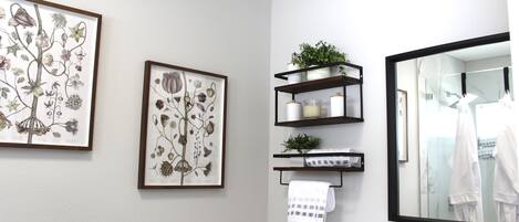 Un baño con impresiones botánicas enmarcadas, un espejo y estantes con artículos de aseo y una planta.