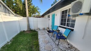 A backyard with a white fence, a small grassy area, a gravel path, and two blue chairs with a potted plant.