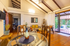 A spacious living area with a high ceiling, a sofa set, a dining table with chairs, and a large window with a view of greenery.
