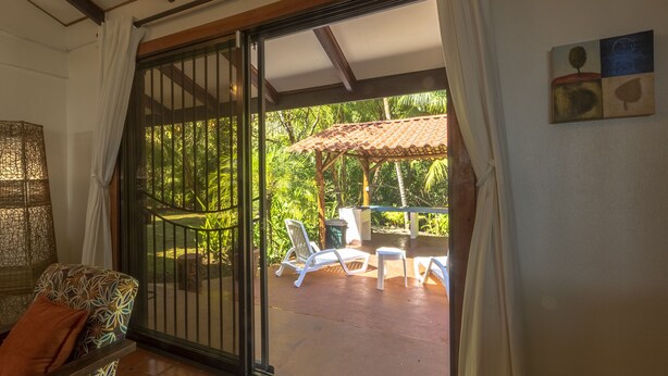 Une pièce dotée d’une porte-fenêtre coulissante donnant sur une terrasse extérieure avec une table et des chaises, le tout entouré d’une végétation luxuriante.