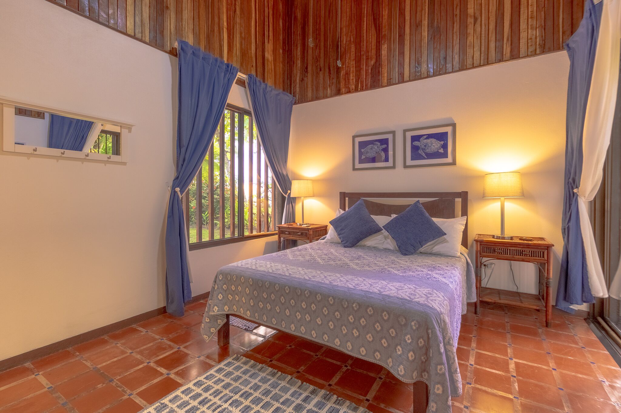 A bedroom with a wooden ceiling, a bed with a patterned bedspread, two wall-mounted lamps, and a window with curtains.