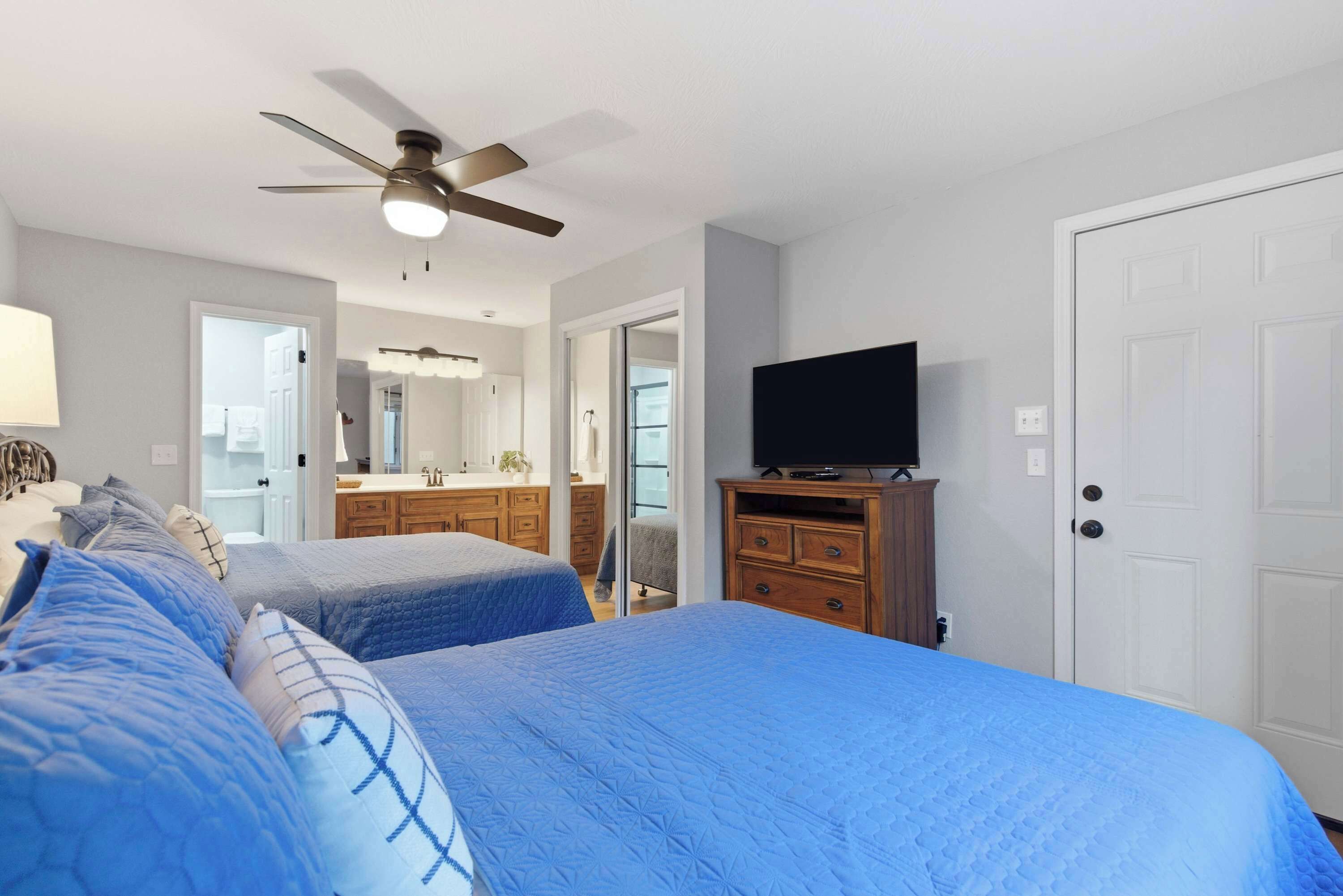 Une chambre avec deux lits, un ventilateur de plafond, un téléviseur posé sur une commode et une porte blanche.