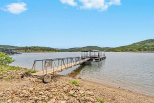 Un muelle a orillas del lago con un paseo de madera que se adentra en el agua, rodeado de terreno rocoso y colinas al fondo.