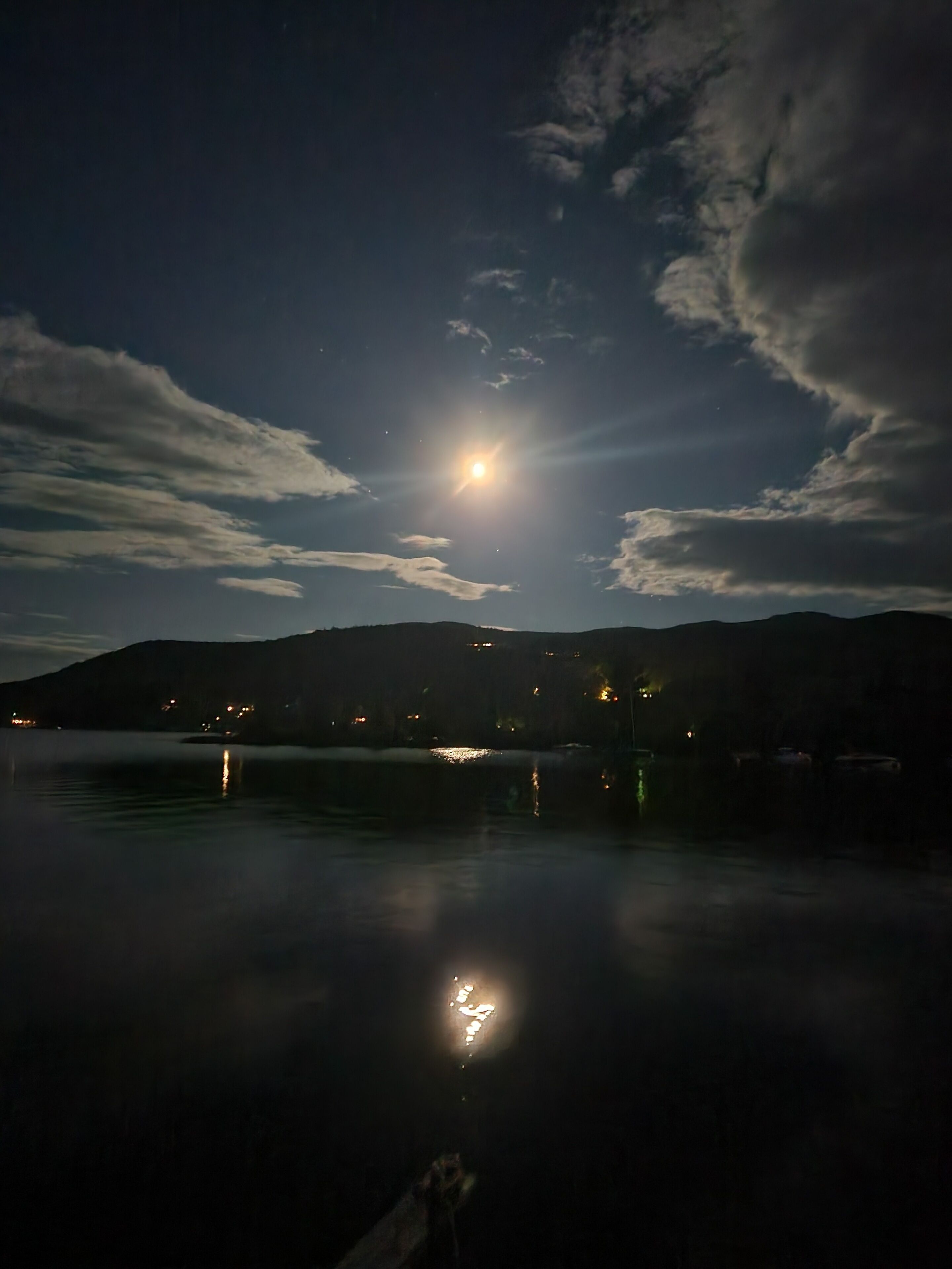A night scene with a moonlit lake, calm water reflecting the moon, and distant hills with scattered lights.