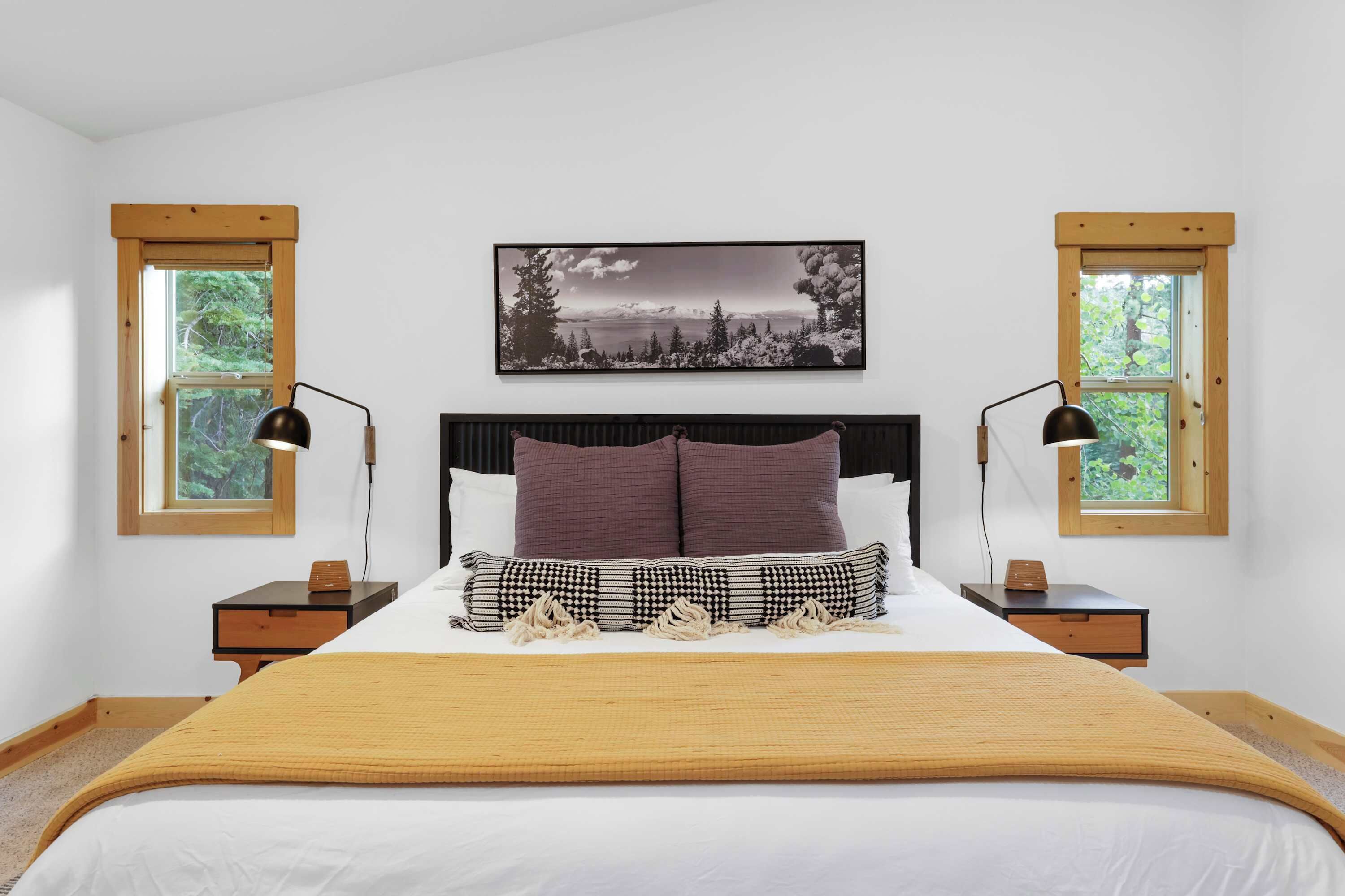 Ein Schlafzimmer mit einem großen Bett, zwei Nachttischen, einem Holzkopfteil, einem gerahmten Bild über dem Bett und zwei Fenstern mit Holzrahmen.