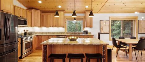 A modern kitchen with wooden cabinets, stainless steel appliances, a large island with bar stools, and a dining area with a large window.