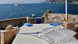 Eine Strandterrasse mit einem Bett, Liegestühlen und Blick auf das Meer und Boote.