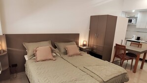Una cama doble con cubrecama acolchado, una mesita de noche con lámpara, un armario, una mesa de comedor con sillas y un microondas al fondo.