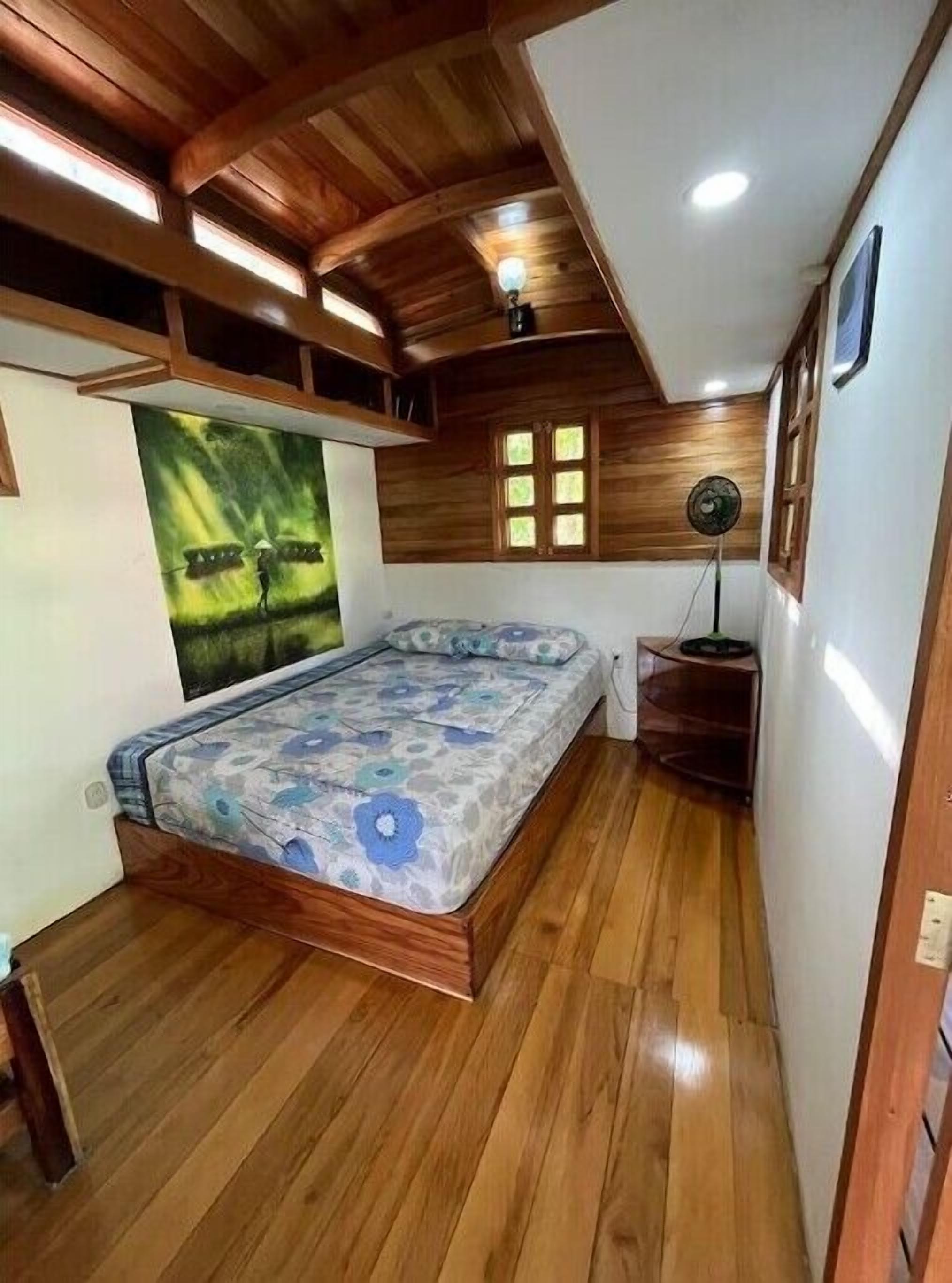 Une chambre avec un plafond en bois, un lit recouvert d’une couvre-lit à motifs floraux, une fenêtre et un ventilateur sur pied.