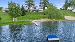 A house with a dock extending into a calm lake, surrounded by lush greenery and trees. - Large Updated Lakefront House on Lake George. Kayaks, games, fire pit, sledding. (West Branch)