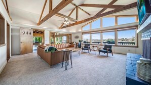 A spacious living room with a high ceiling, exposed wooden beams, a large window offering a view, and a cozy seating area. - Large Updated Lakefront House on Lake George. Kayaks, games, fire pit, sledding. (West Branch)