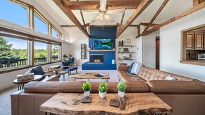 A spacious living room with a large sectional sofa, a wooden coffee table, and a fireplace. There are multiple windows providing natural light and a view of the outdoors. - Large Updated Lakefront House on Lake George. Kayaks, games, fire pit, sledding. (West Branch)